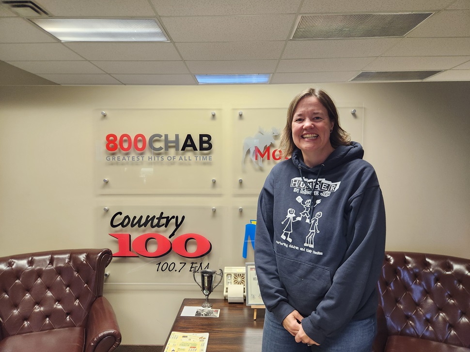 Sharla standing in front of the Golden West Moose Jaw logos wearing a Hunger in Moose Jaw sweater.