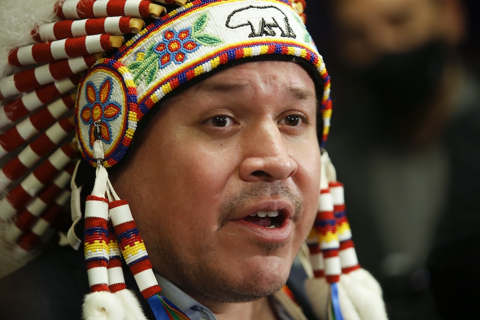 A closeup of a man speaking. He wears a large Indigenous head dress with white feathers, and colourful beading