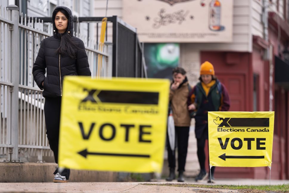 Small yellow signs that say "Elections Canada - VOTE" with black arros line a sidewalk as 3 people walk in the photo