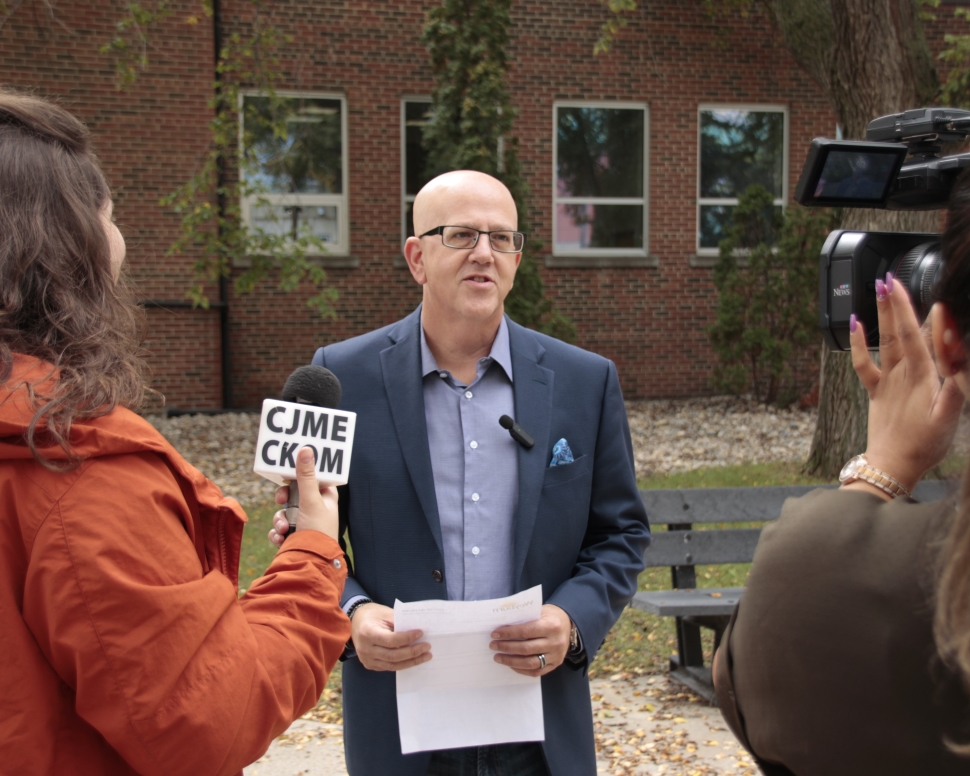 A man in a blueish-grey blazer and a blue shirt in front of members of the media
