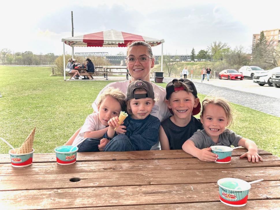 A mom and 4 kids sit with ice cream cones at a picnic table and smile