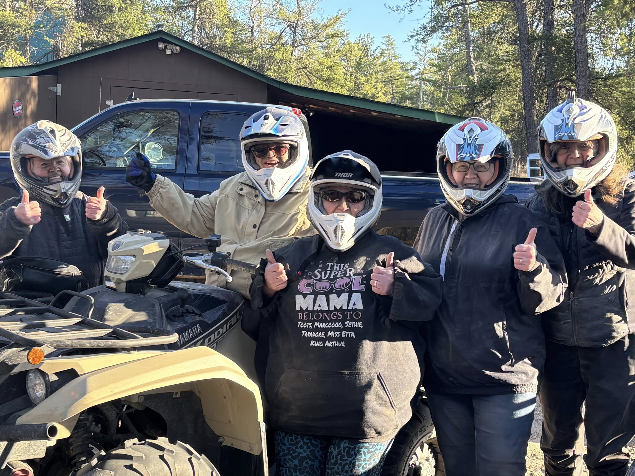 5 people stand around an ATV wearing helmets and a bit dirty from quadding - they all give thumbs up to the camera