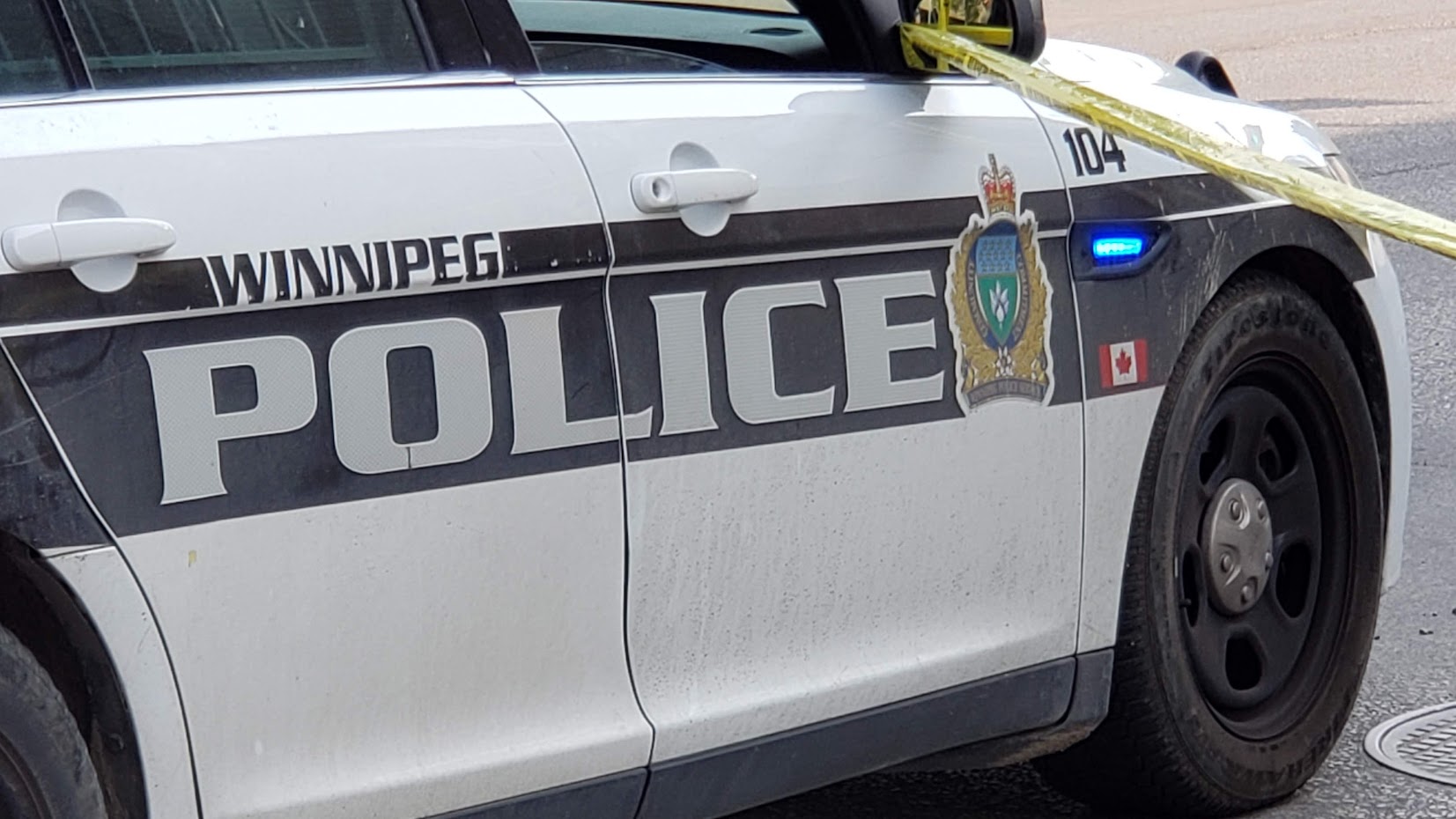 A close-up of the side of a white police cruiser with a black horizontal stripe down the side. "Winnipeg Police" is in letters with a WPS crest. Yellow police tape is attached to the mirror of the car and runs out of frame