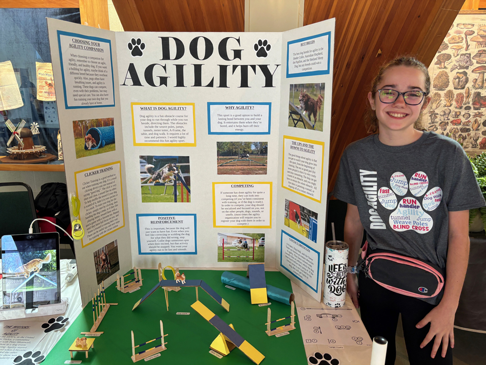 A female middle-school student stands beside her agility dog training display.