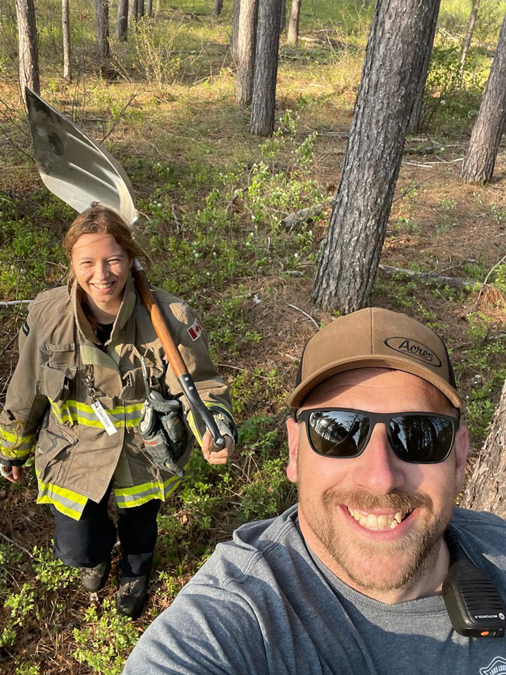 Two firefighters, a man and a woman, in a forest, carrying a shovel.