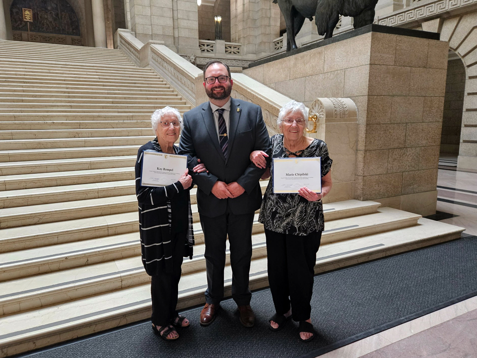 A man stands between two elderly women, they are holding up certificates.