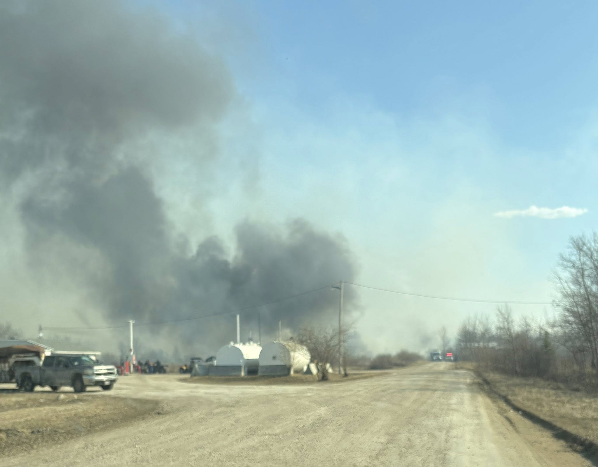 Emergency Response at Rural Oil Storage Site: Thick Smoke Billows Over Unpaved Road and Industrial Fuel Tanks