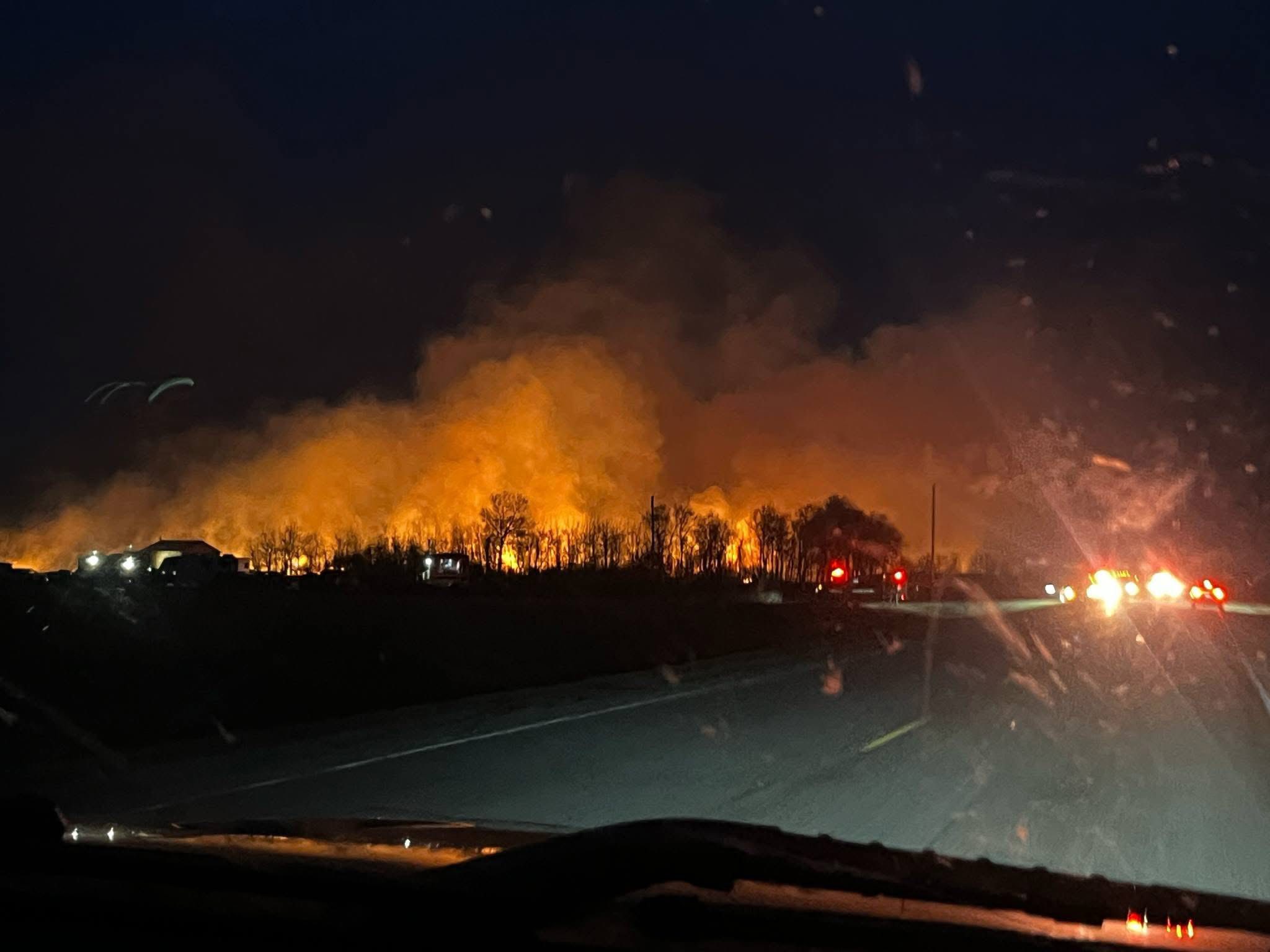 Nighttime Wildfire on Rural Highway: Emergency Crews Battle Intense Orange Blaze and Thick Smoke Along Tree Line at Country Road Frontage