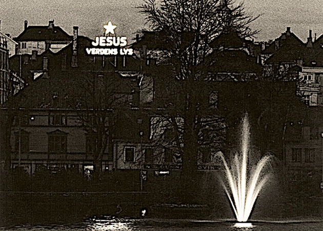 A black and white photo at night shows a lit up sign with a star and the words "Jesus verdens lys"