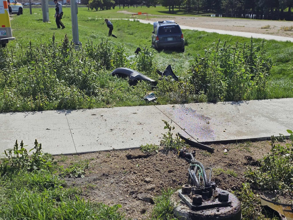 SUV sits in a grassy ditch at the side of Saskatchewan Avenue West, with debris scattered and a damaged traffic light base in the foreground. Emergency personnel are visible in the background.