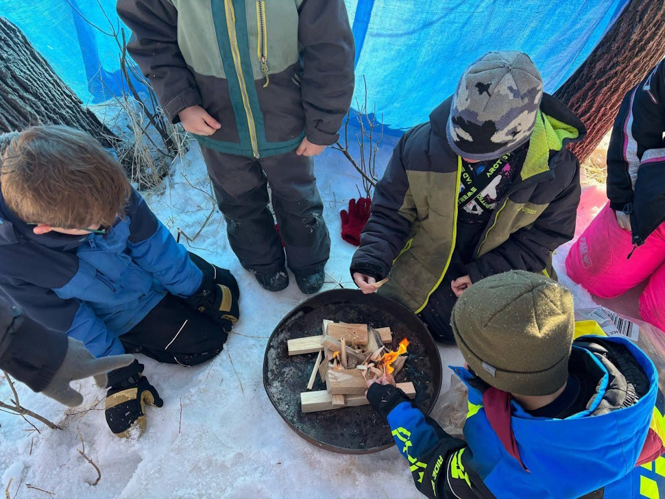 A group of Scouts gathered around a campfire in a snowy winter setting, bundled in warm clothing.