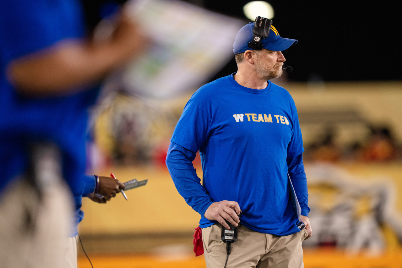 A football coach watches from the sidelines. He wears a headset and has a hand on an audio receiver clipped to his belt