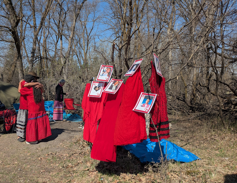 Deb Parker and Jeanna Emms hugging before the second annual Red Dress Day Walk next to a display of red dresses and photos, honouring their missing and murdered loved ones