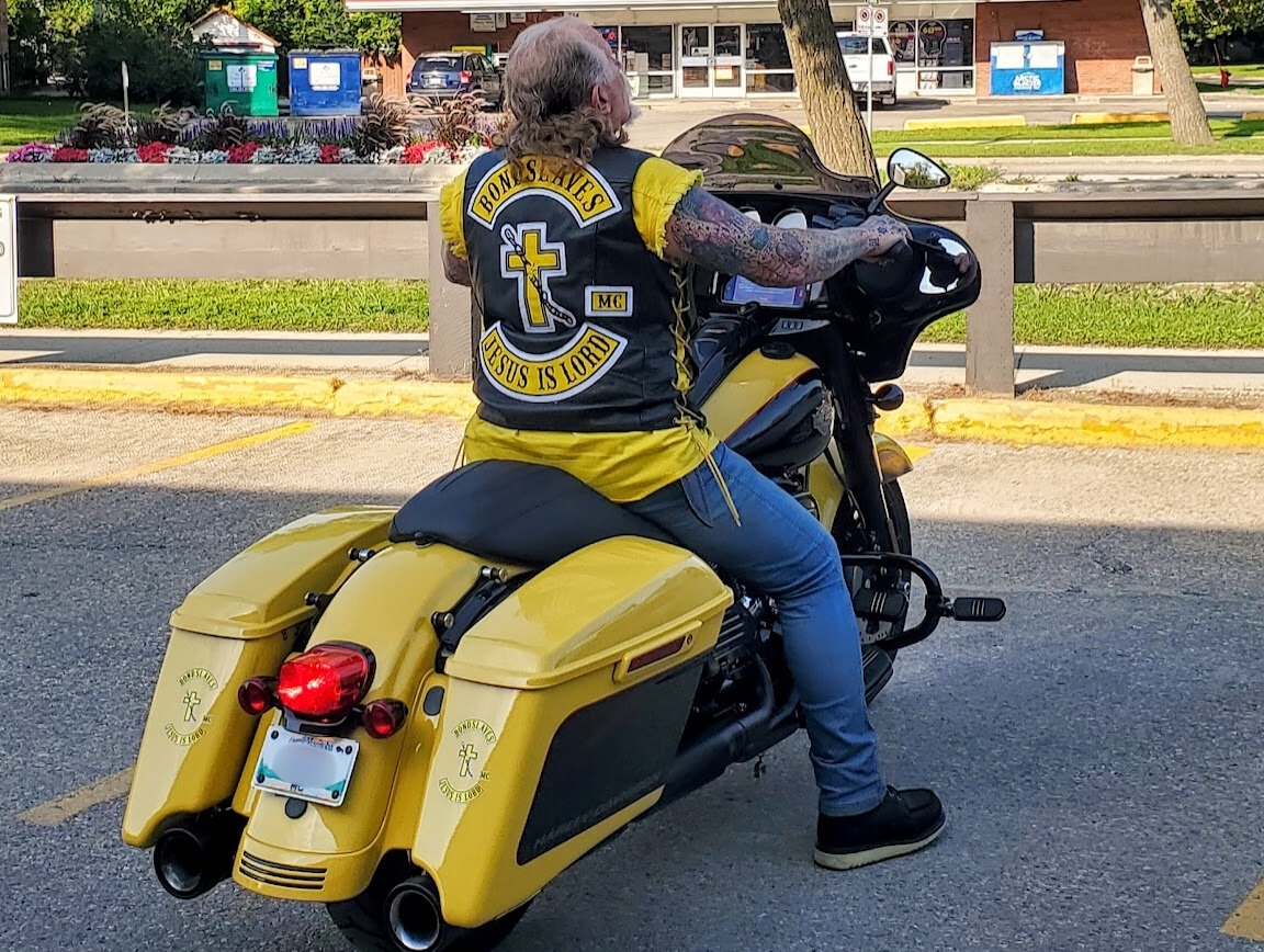 A shot from behind of a man sitting on a black and yellow Harley Davidson. He wears a black vest with a black and yellow 3-piece patch. The top rocker says "Bondslaves," the bottom rocker says "Jesus Is Lord" and the centre piece is a cross with a chain running across that is broken.
