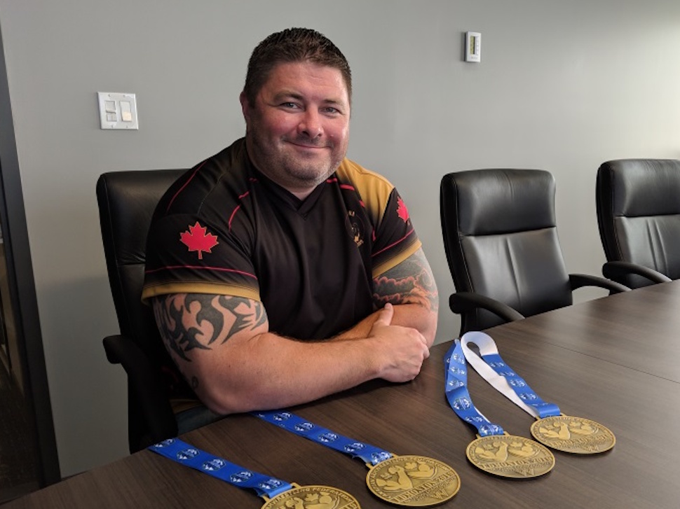 Ryan Espey smiling and posing with his collection of arm wrestling medals displayed on a table.