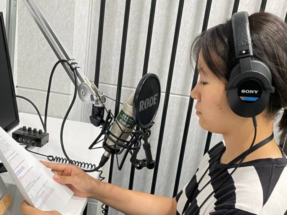 A woman sits in a recording booth. She wears a pair of sony over-ear headphones and sits behind a mic with pop filter and holds a script