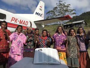 A group of people stand behind a large Bible in their own language, and in front of a plane