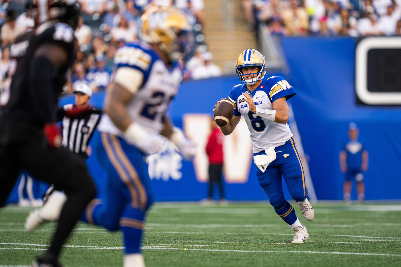 A football quarterback runs with the ball and preparest to throw it. A player runs through the frame in the foreground