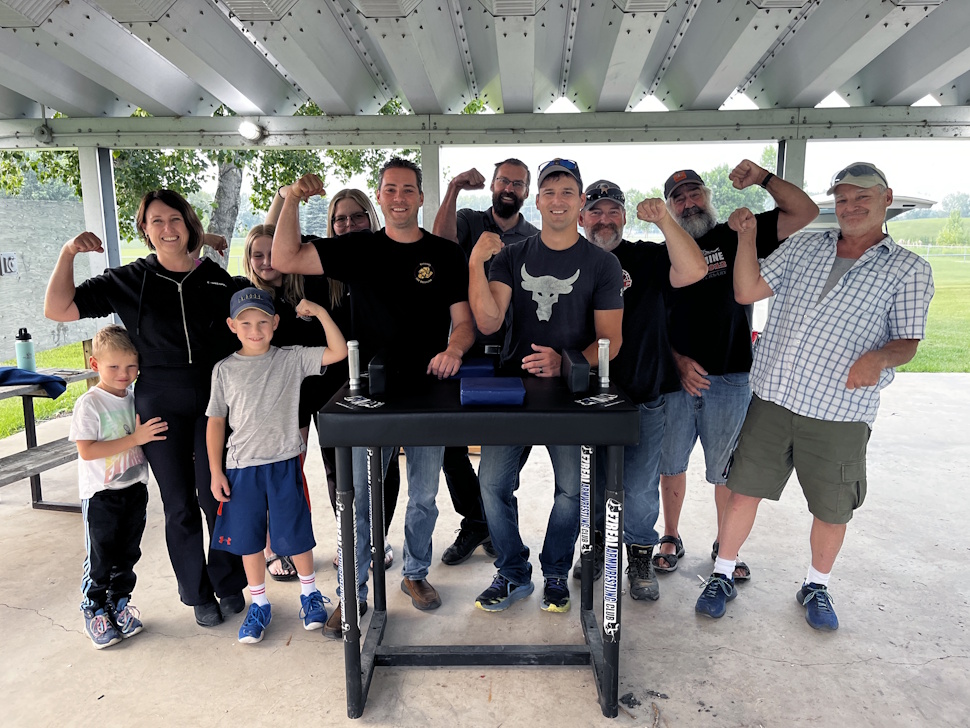 Members of the Portage Arm Wrestling Club flex their muscles and pose together outdoors at Rotary Republic of Manitobah Park on a summer evening.