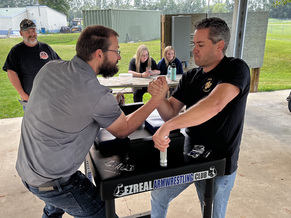Two men engaged in an intense arm wrestling match, with Ryan Heron on the right, focused and exerting effort.