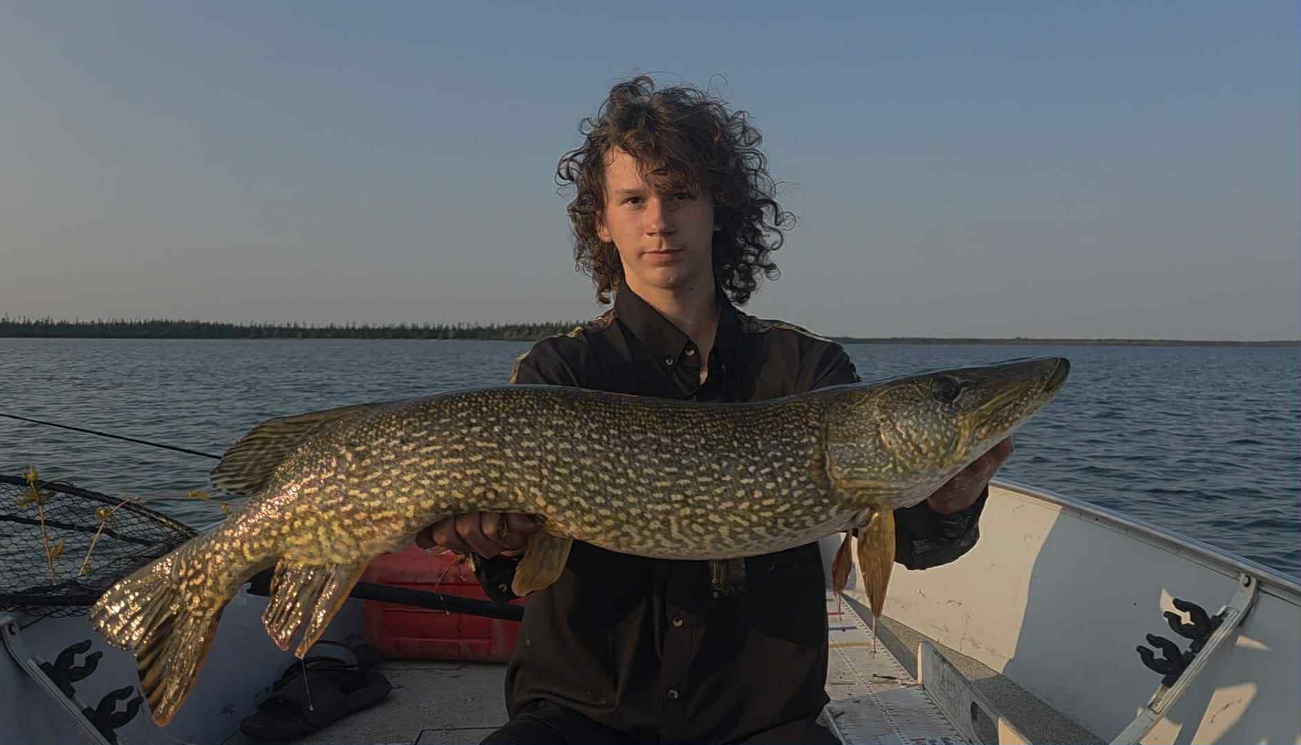 A young man, Paul, holding a large, freshly caught pike horizontally with both hands. He is looking directly at the camera with a calm expression. He has long, curly dark hair and is wearing a black shirt. He is sitting in a boat on a calm body of water with a shoreline and trees visible in the distance under a clear sky.