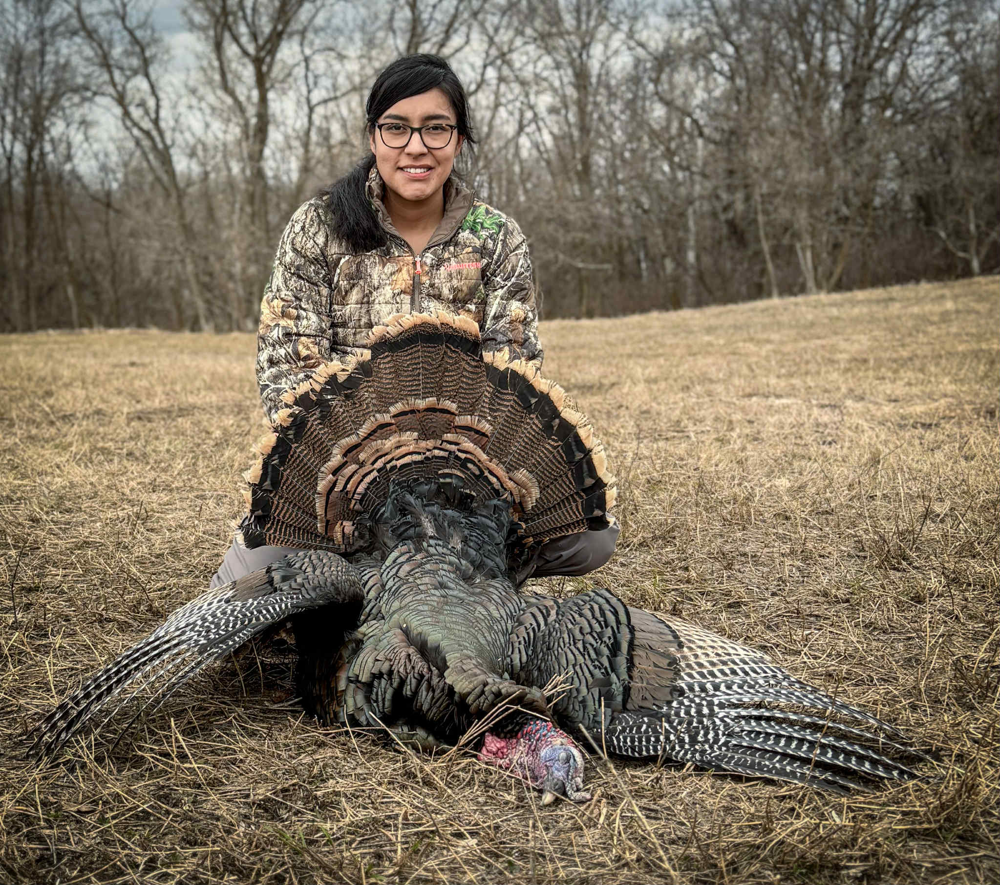 A medium-shot horizontal image of a young woman, Ruth Spence, kneeling behind a large, deceased turkey with its tail feathers fanned out. She is smiling and wearing a camouflage jacket and glasses. The ground is covered in dry grass, and there are bare trees in the background.