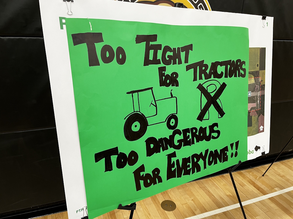 A handmade protest sign that says “Too tight for tractors, too dangerous for everyone,” taped over a government posterboard at the Carberry Collegiate gymnasium during the June 25 meeting.
