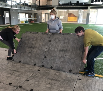 Three people lift large square tiles of hard floor covering the turf