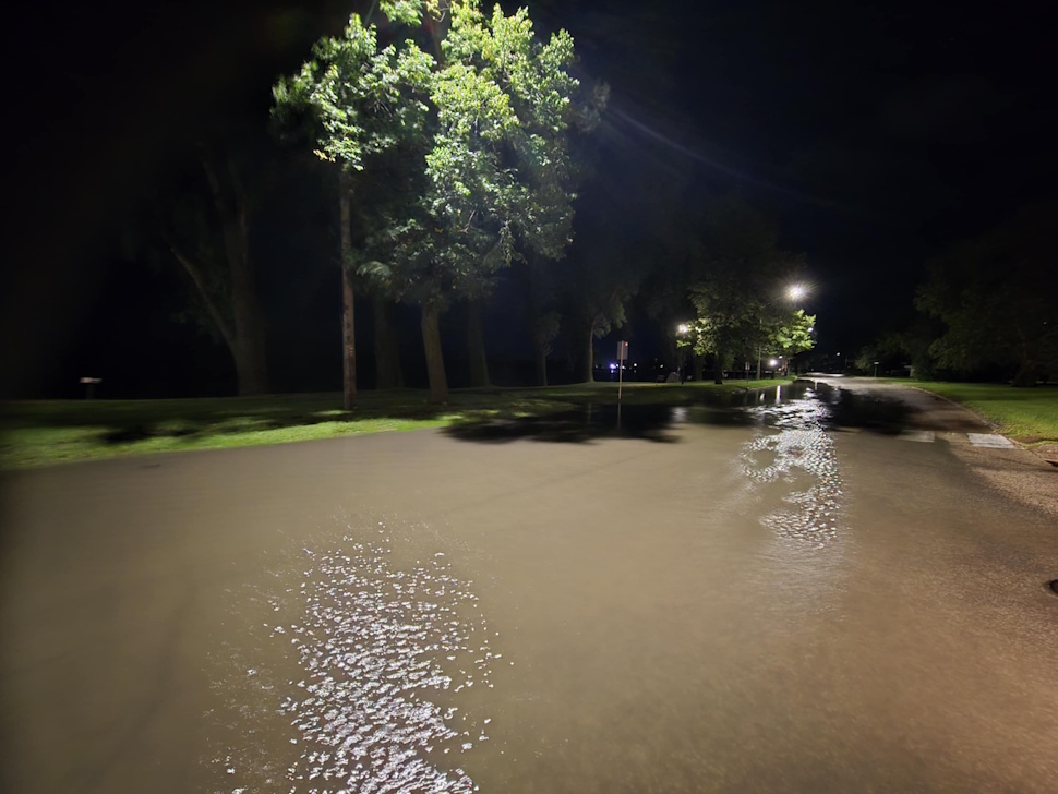 Crescent Road in Portage la Prairie submerged in floodwaters after heavy overnight rain, with vehicles navigating the water-covered street.