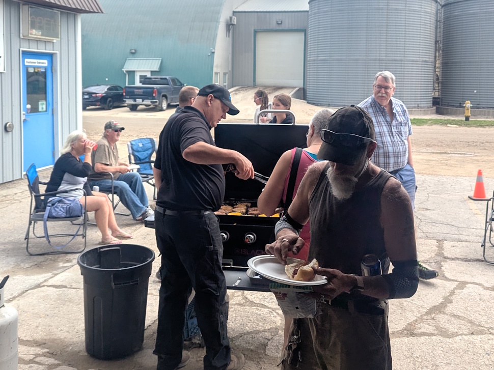 People gathered outdoors enjoying BBQ food at a fundraiser event, with plates of burgers, hot dogs, and drinks visible.
