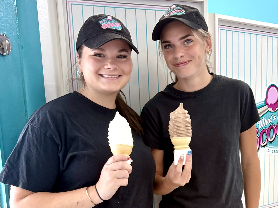 Two young female employees smile while holding soft serve ice cream cones inside an ice cream shop.