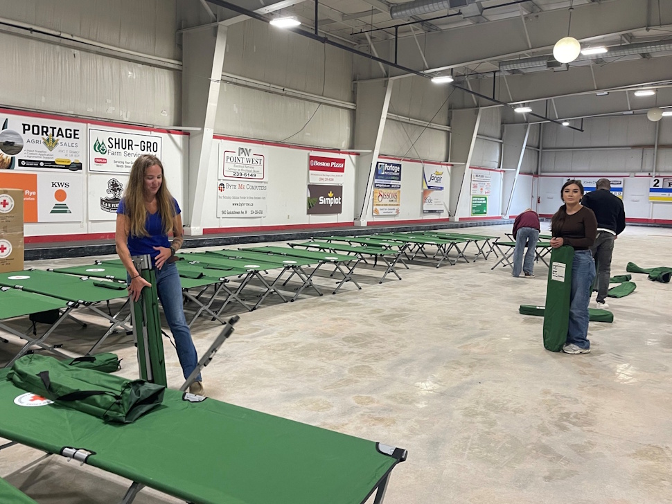 Group of young women and staff setting up rows of cots inside the Portage Curling Club for incoming wildfire evacuees.
