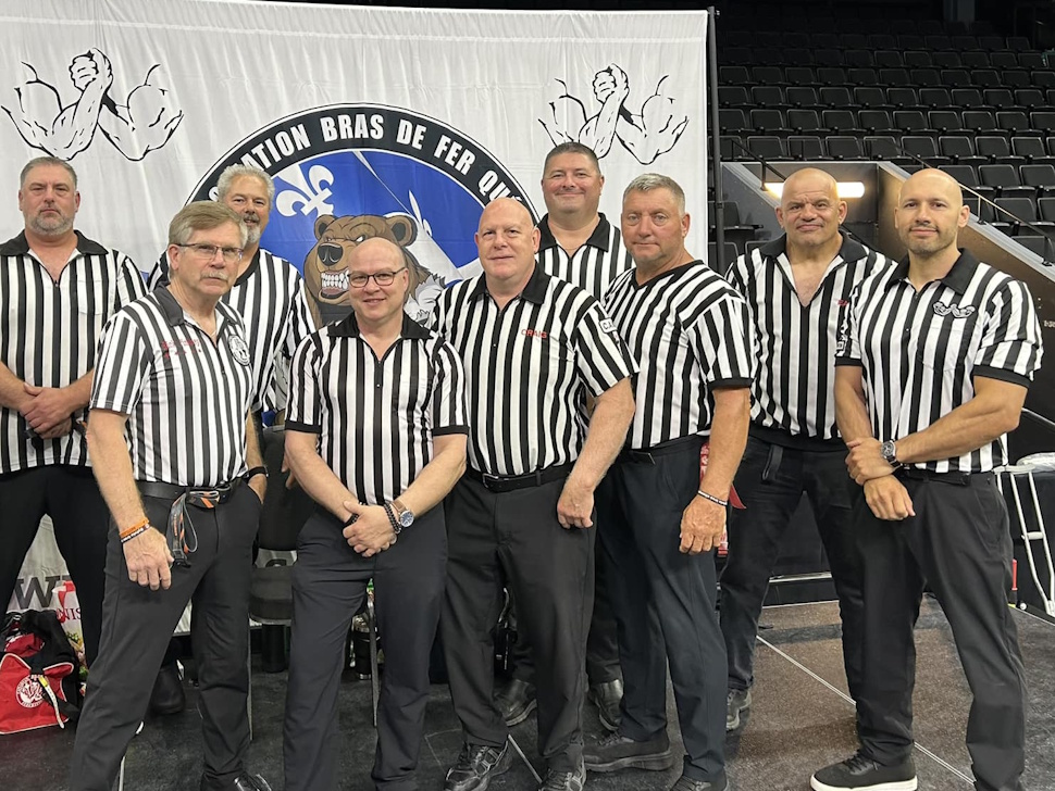 Ryan Espey stands in referee uniform, focused, while officiating an arm wrestling match at the Quebec event.