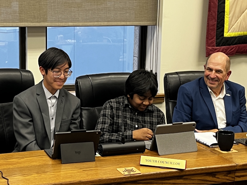 Two youth councillors sit at a council table using tablets, with Portage councillor Ferron Nichols seated to the right, smiling. François Barnachea is on the far left, dressed in a suit.